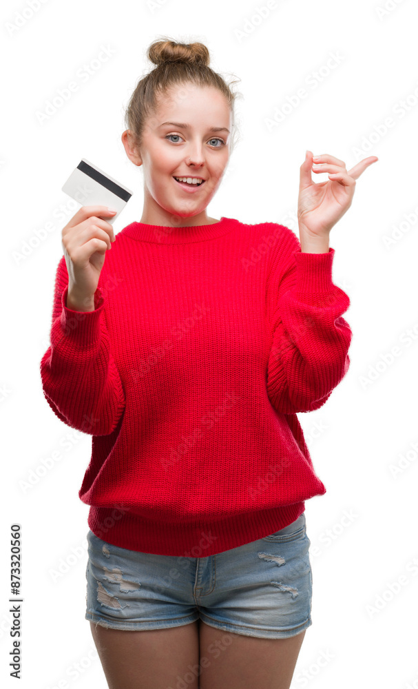 Young blonde woman holding credit card very happy pointing with hand and finger to the side Young blonde woman holding credit card very happy pointing with hand and finger to the side