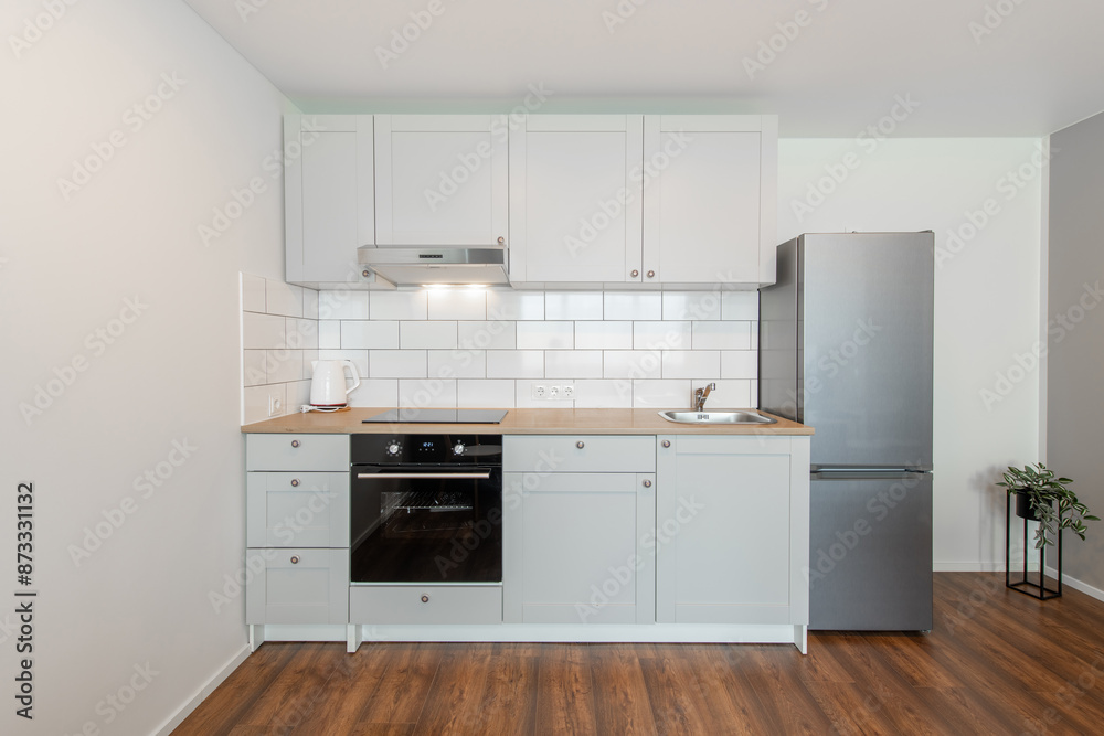 Scandinavian Style Cozy Modern Open Bright Minimalism Kitchen Home Interior. Oven, Faucet, Kettle, Fridge and Furniture in Background. Dark Brown Flooring. Flower. White Cabinets Scandinavian Style Cozy Modern Open Bright Minimalism Kitchen Home Interior. Oven, Faucet, Kettle, Fridge and Furniture in Background. Dark Brown Flooring. Flower. White Cabinets