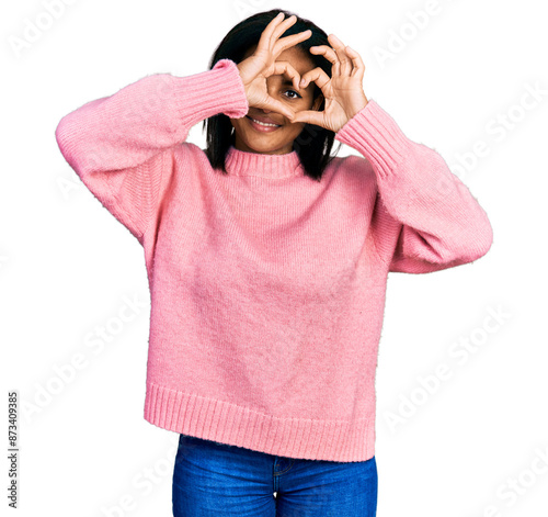 Beautiful hispanic woman wearing casual winter sweater doing heart shape with hand and fingers smiling looking through sign