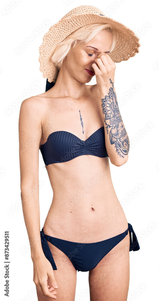 Young blonde woman with tattoo wearing bikini and summer hat tired rubbing nose and eyes feeling fatigue and headache. stress and frustration concept. Young blonde woman with tattoo wearing bikini and summer hat tired rubbing nose and eyes feeling fatigue and headache. stress and frustration concept.