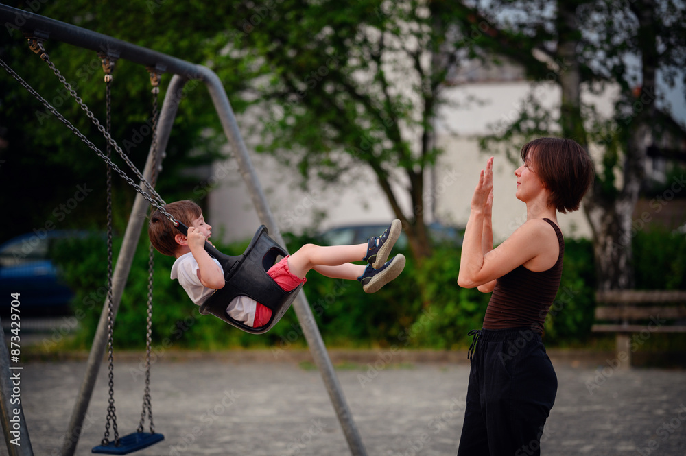 Obraz premium A mother pushing her laughing young son on a swing at the playground, capturing a moment of pure joy and playful bonding together outdoors.