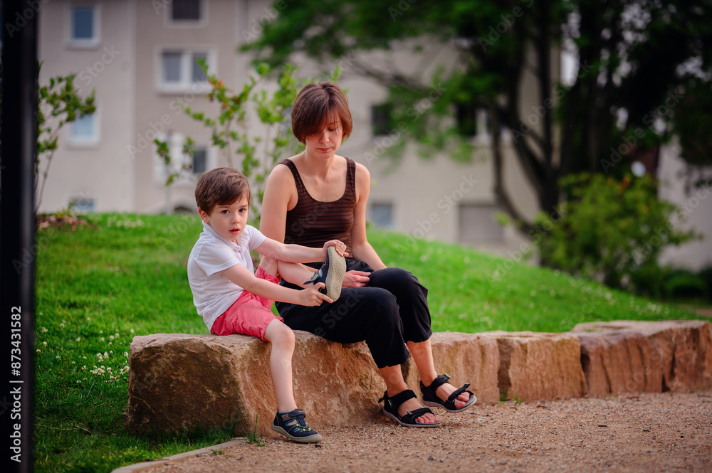 A mother and her young son sit on a large rock in a park. The boy examines his shoe while the mother watches him calmly. The scene captures a quiet, reflective moment in the outdoors. A mother and her young son sit on a large rock in a park. The boy examines his shoe while the mother watches him calmly. The scene captures a quiet, reflective moment in the outdoors.