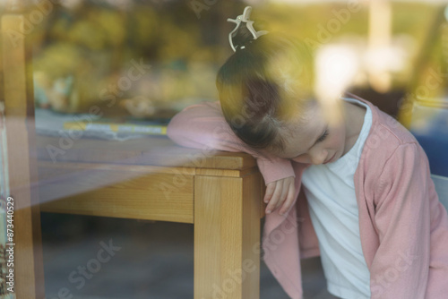 Tired little girl in a pink blouse sits at a wooden table by a large window, head bowed. Indoor portrait with soft light, quiet mood, childhood struggle and emotional concept.