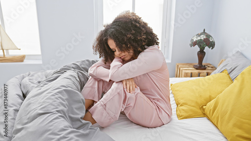 Canvastavla A young hispanic woman with curly hair feels saddened while sitting in a cozy bedroom setting, conveying a mood of contemplation or solitude