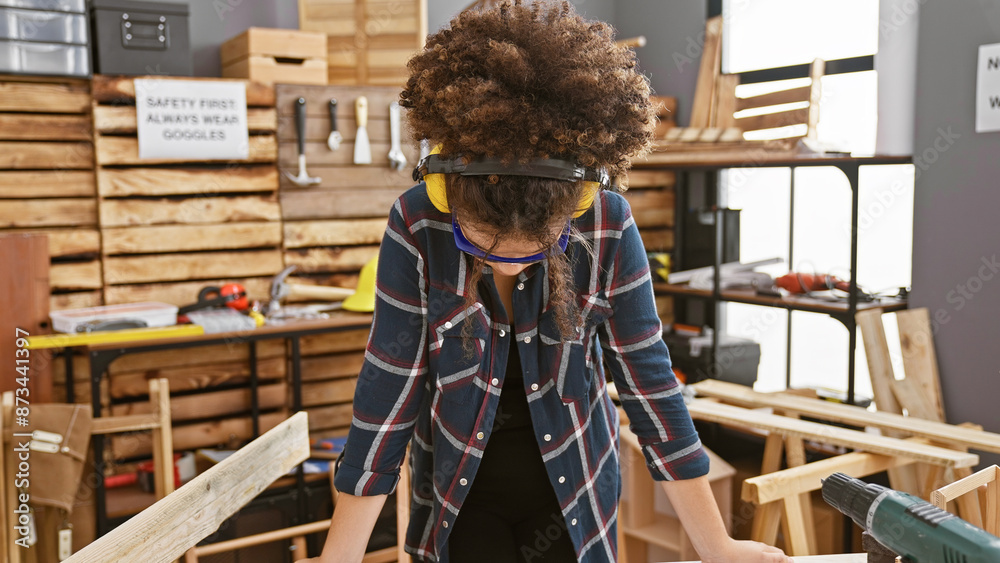 Hispanic woman with curly hair wearing safety glasses works in a carpentry workshop. Hispanic woman with curly hair wearing safety glasses works in a carpentry workshop.
