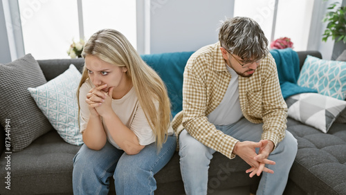 A worried couple sits apart on a couch in a modern living room, showing tension in their relationship. photo