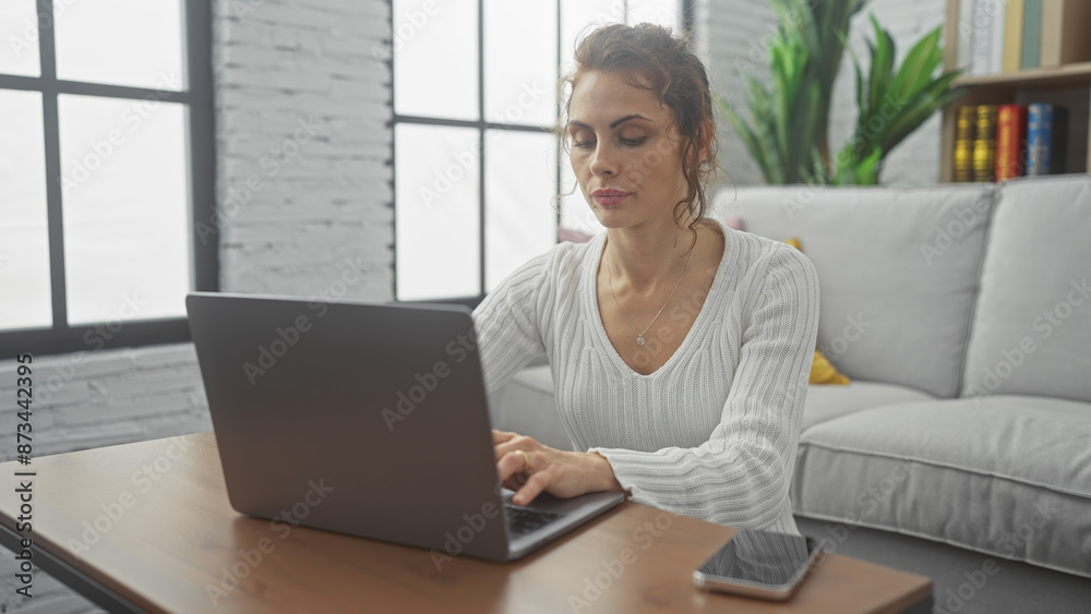 A focused young woman using a laptop in a modern living room with natural light and minimalistic decor. A focused young woman using a laptop in a modern living room with natural light and minimalistic decor.