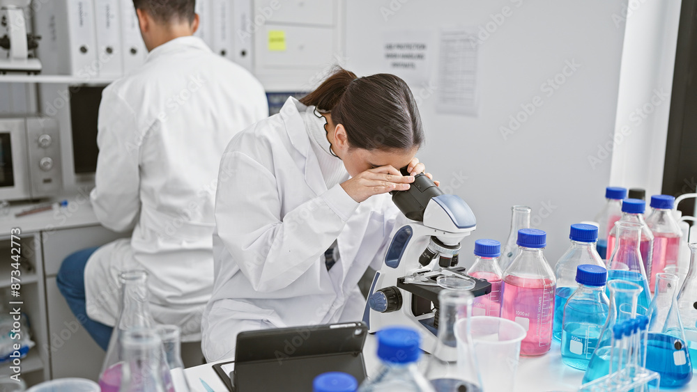 A woman scientist using a microscope in a laboratory with a male coworker and various chemical bottles in the background. A woman scientist using a microscope in a laboratory with a male coworker and various chemical bottles in the background.