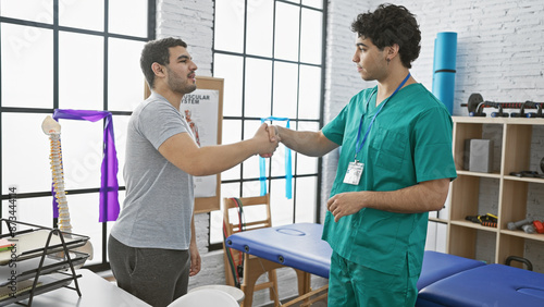 In a bright physiotherapy room, a male therapist wearing scrubs and his male patient handshake, surrounded by rehabilitation equipment. © Krakenimages.com