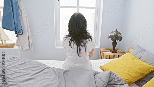 A woman sits contemplatively on a bed in a bright bedroom, facing a window, surrounded by modern home decor.