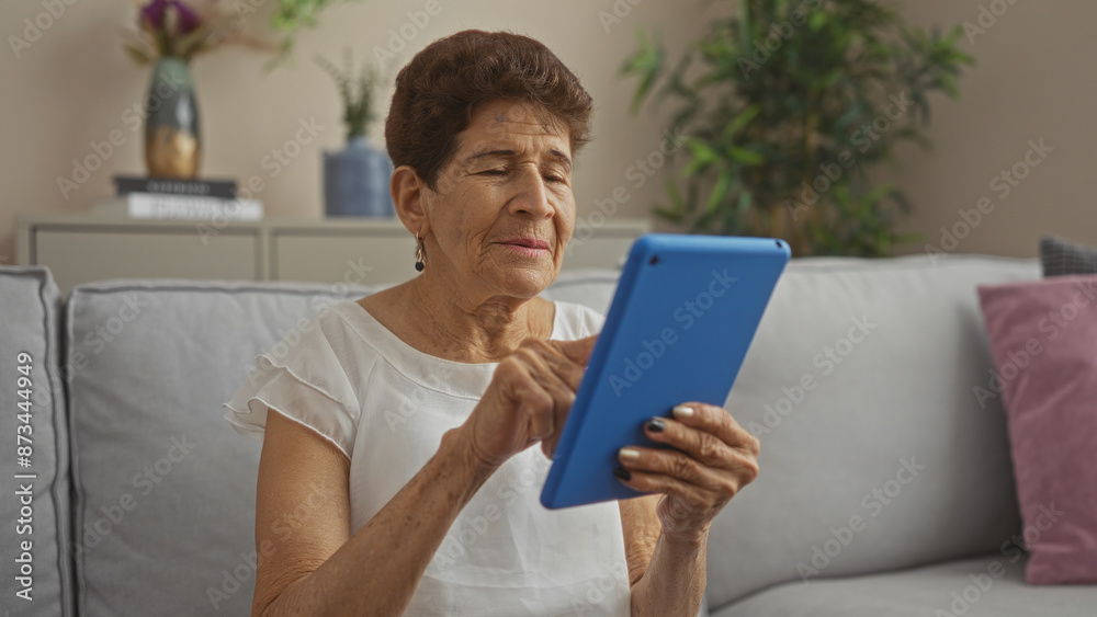 Elderly hispanic woman with short hair using a blue tablet indoors on a sofa, in a well-decorated living room. Elderly hispanic woman with short hair using a blue tablet indoors on a sofa, in a well-decorated living room.