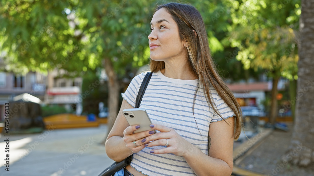 A thoughtful young woman with a smartphone relaxes in a sunny park filled with green trees. A thoughtful young woman with a smartphone relaxes in a sunny park filled with green trees.
