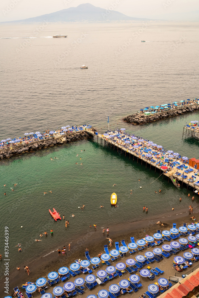 Sorrento, Italy - 07.24.2019: People relaxing on the beach on the pier and swimming in the sea on inflatable air beds in Sorrento and boats floating against mount vesuvius volcano