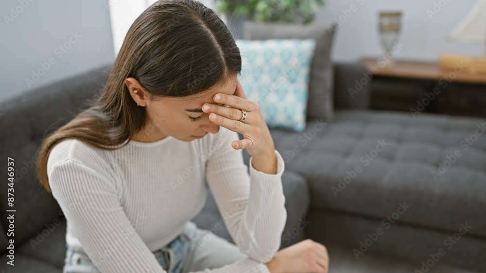 A stressed young woman indoors touches her forehead, showcasing worry or a headache in a comfortable living room. A stressed young woman indoors touches her forehead, showcasing worry or a headache in a comfortable living room.
