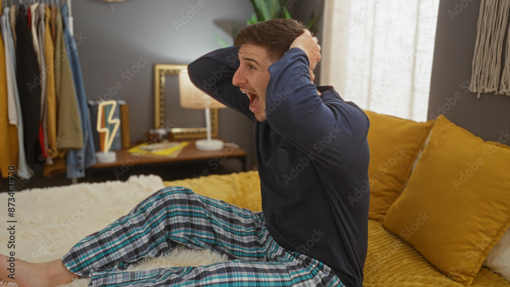 Fototapeta premium A surprised young hispanic man sits on a bed with his hands on his head, in a cozy bedroom with warm lighting, surrounded by decor and clothes in the background.