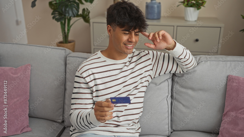 Handsome young hispanic man smiling while holding a credit card in a cozy living room with indoor plants and modern decor. Handsome young hispanic man smiling while holding a credit card in a cozy living room with indoor plants and modern decor.