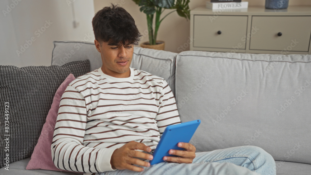 A handsome young hispanic man enjoys a tablet in his cozy living room, seated comfortably on a modern sofa, highlighting the relaxed indoor ambiance of his stylish home. A handsome young hispanic man enjoys a tablet in his cozy living room, seated comfortably on a modern sofa, highlighting the relaxed indoor ambiance of his stylish home.
