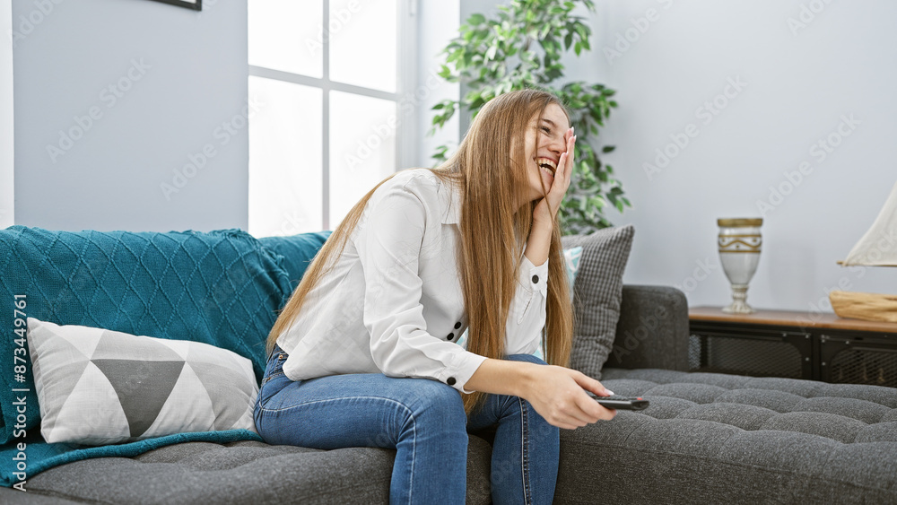 Laughing young blonde woman holding a remote in a cozy living room. Laughing young blonde woman holding a remote in a cozy living room.