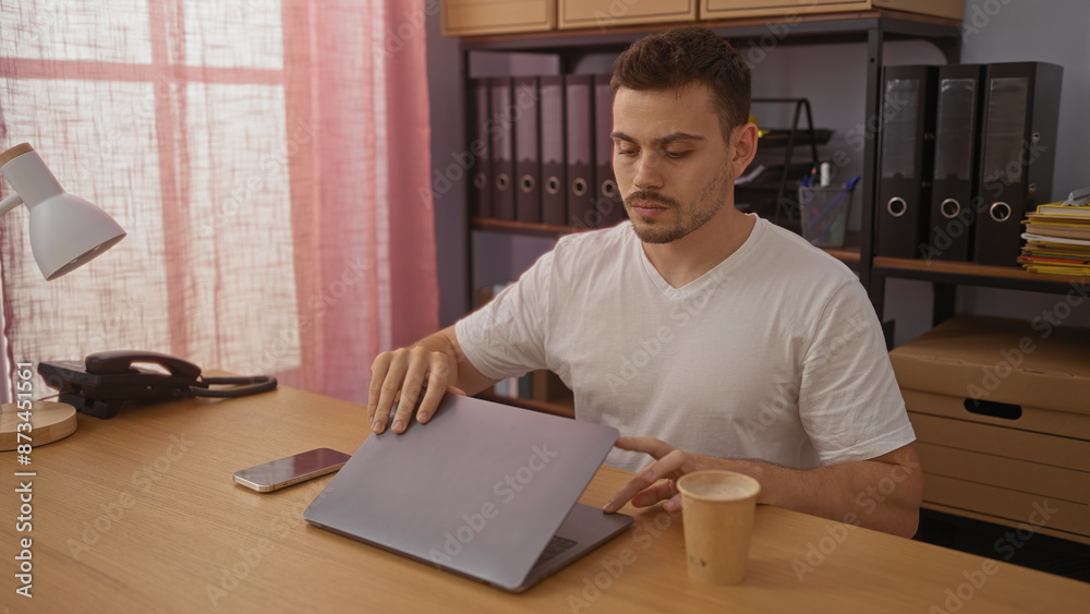 A handsome young hispanic man with a beard, wearing a white shirt, working on a laptop in a modern office with pink curtains and organized shelves in the background. A handsome young hispanic man with a beard, wearing a white shirt, working on a laptop in a modern office with pink curtains and organized shelves in the background.