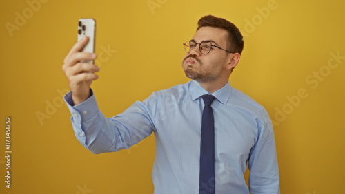 A handsome young hispanic man in a blue shirt and tie takes a selfie against a vibrant yellow background.