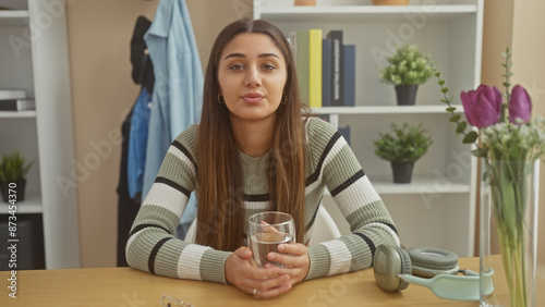 A young woman sits at a table indoors, looking thoughtful, surrounded by plants and modern decor.