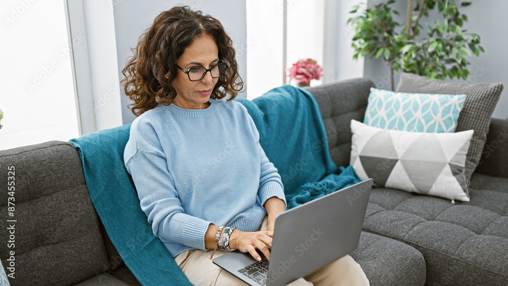 Hispanic woman in blue sweater working on laptop indoors on gray sofa with teal throw. Hispanic woman in blue sweater working on laptop indoors on gray sofa with teal throw.
