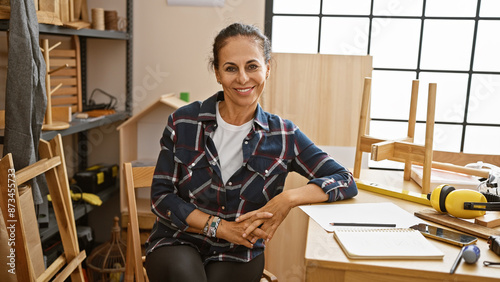 Obraz na płótnie Smiling woman in a plaid shirt sitting in a sunny carpentry workshop surrounded by woodwork tools