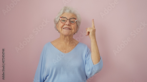 Принти на полотні A cheerful senior woman with glasses pointing upwards against a pink isolated background