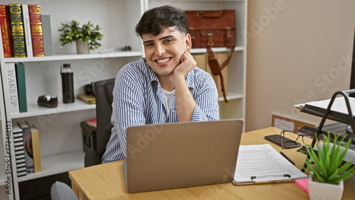 A young adult man smiles while working on a laptop in a modern office setting with books and plants.