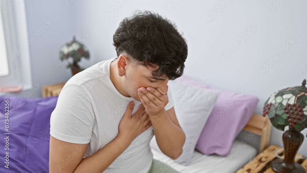 A distressed young man holding his chest and covering his mouth while sitting on a bed in a bedroom. A distressed young man holding his chest and covering his mouth while sitting on a bed in a bedroom.