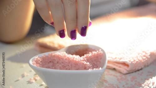 Female hand sprinkling pink sea salt in a dish in a beauty studio, showcasing new purple manicure, concept of femininity and manicure