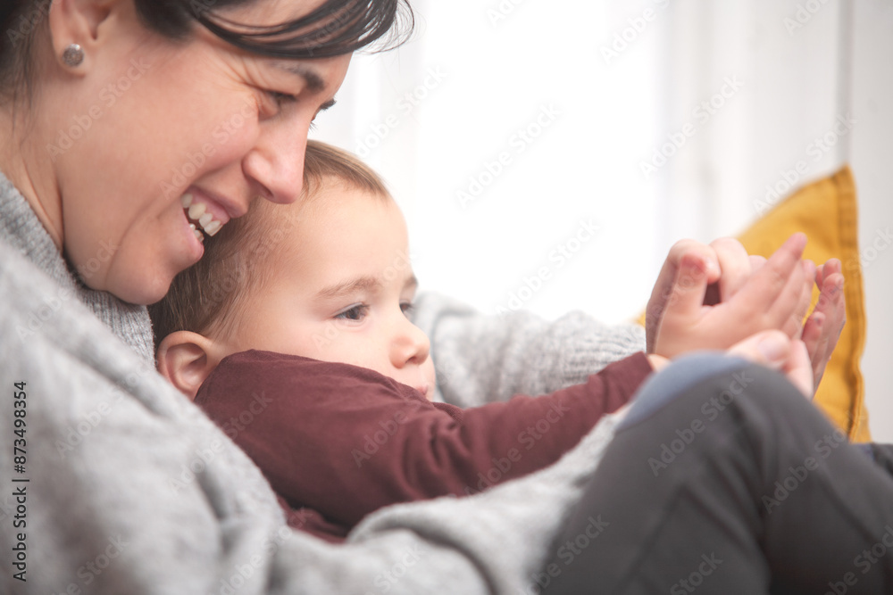 Close-up of toddler and his mother sitting together hugging smiling while playing with their hands. Motherhood and family. Close-up of toddler and his mother sitting together hugging smiling while playing with their hands. Motherhood and family.