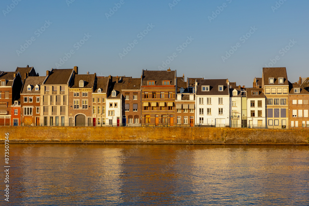 Fototapeta premium Summer cityscape of architecture traditional houses in Wyck district with warm sunlight in the afternoon, The Meuse (Maas) riverside view under blue sky in evening, Maastricht, Limburg, Netherlands.