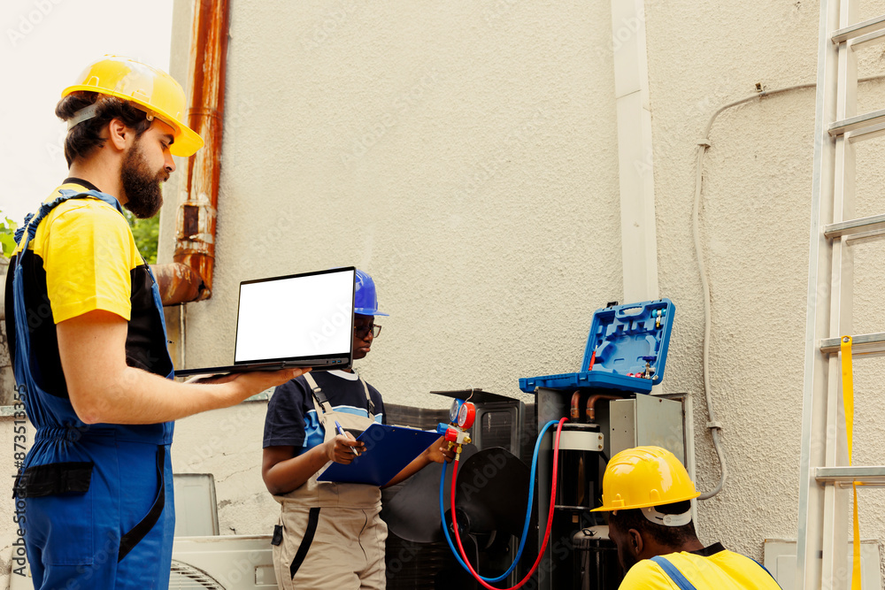 Proficient worker cleaning outdoor air conditioner internal coolants, checking refrigerant level and calibrating thermostats while diverse teamworking colleagues check maintenance procedures on laptop Proficient worker cleaning outdoor air conditioner internal coolants, checking refrigerant level and calibrating thermostats while diverse teamworking colleagues check maintenance procedures on laptop