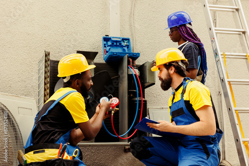 Precise servicemen expertly refilling refrigerant in outside air conditioner while using manifold indicators to reliably measure the pressure in HVAC system, ensuring proper cooling performance photo