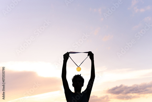 Silhouette of a male athlete raising his gold medal to celebrate his victory in a major sports event. In the background, there is a beautiful sky.