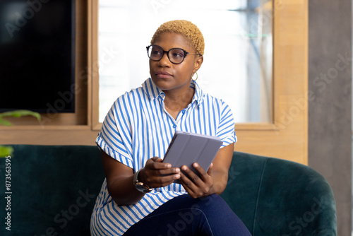 Holding tablet, african american woman sitting on sofa and looking away thoughtfully