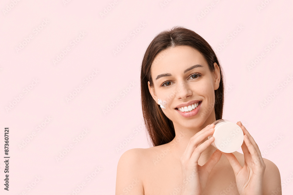 Beautiful young woman with cream on cheek holding jar of sunscreen against pink background Beautiful young woman with cream on cheek holding jar of sunscreen against pink background