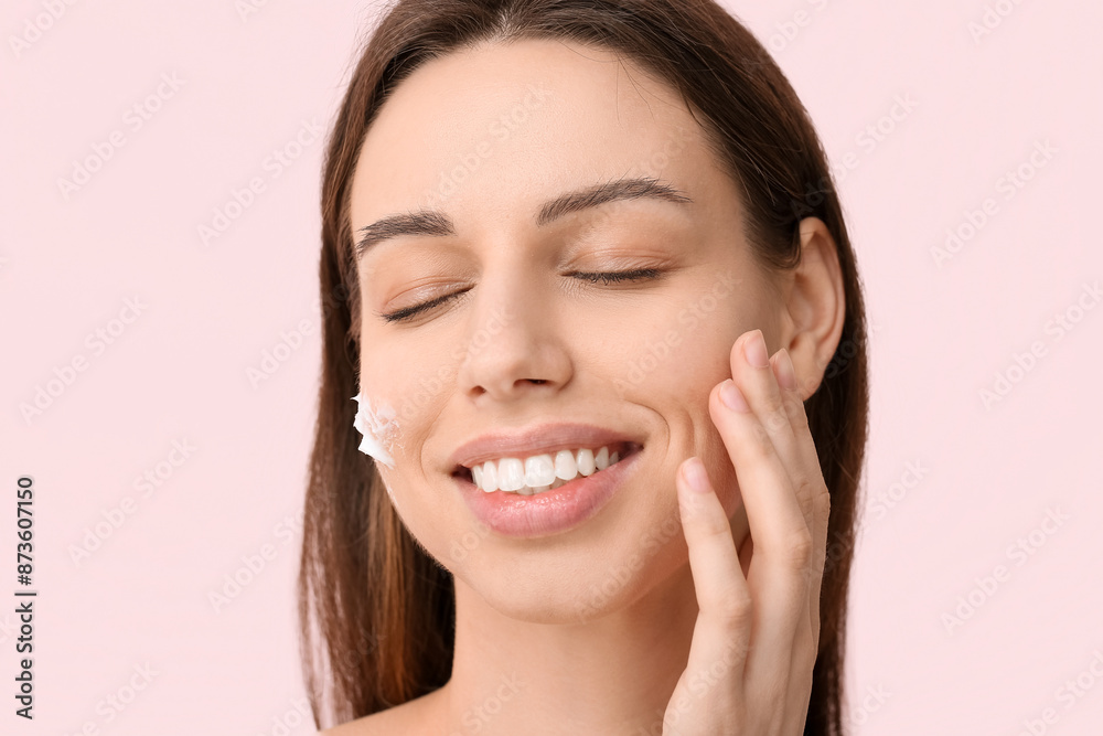 Beautiful young woman with cream on cheek against pink background