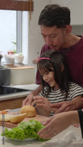 Young Asian Father And Mother Teaching Their Daughter Cutting Bread On Cutting Board To Make Burger At Home