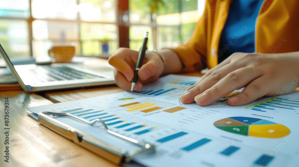 Person analyzing financial charts and graphs with a pen, working on a report at a desk with a laptop. Person analyzing financial charts and graphs with a pen, working on a report at a desk with a laptop.