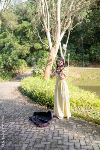Tableau sur toile Young Chinese Malaysian woman in her 20s playing violin near a fountain pond in a large park in Bukit Jalik, Malaysia, wearing a yellow dress