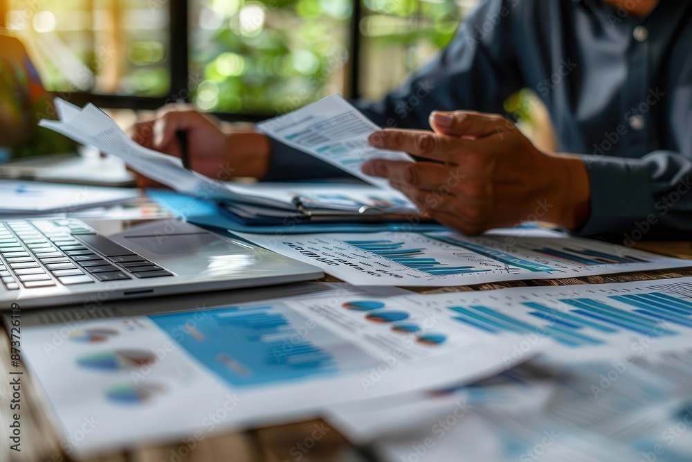 Business professional analyzing financial graphs and charts, working on a laptop in a modern office setting with papers and documents. Business professional analyzing financial graphs and charts, working on a laptop in a modern office setting with papers and documents.
