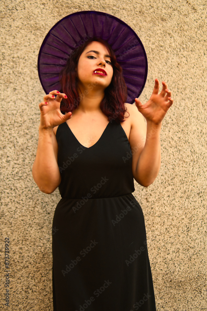 Fototapeta premium beautiful woman with makeup, wearing dress and witch hat, looking and with vampire teeth, a concrete background in an outdoor park. halloween party or celebration