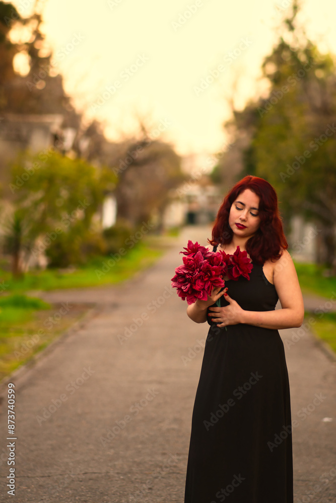 Fototapeta premium Beautiful woman with long black dress, holding bouquet of red flowers, in a cemetery, sad and melancholic look. in an outdoor forest. Halloween party and celebration.