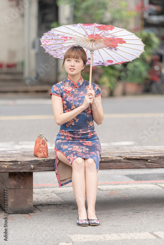 Young Taiwanese woman in her 20s wearing blue cheongsam sitting on a bench along a street in Wanhua District, Taipei, Taiwan, pointing at a Chinese-patterned umbrella. A blend of tradition and modern photo