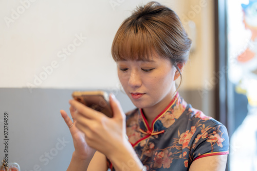 Young Taiwanese woman in her 20s wearing a blue cheongsam, sitting at a table in a Chinese restaurant in downtown Wanhua District, Taipei, Taiwan, using her smartphone while waiting for her ordered fo photo