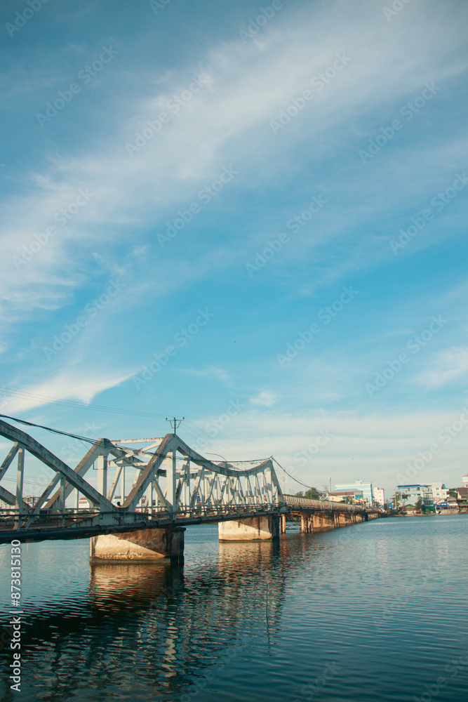 Naklejka premium A calm river flows under a the famous old bridge in Kampot town in Cambodia, reflecting the clear blue sky and scattered clouds, with a cityscape in the background