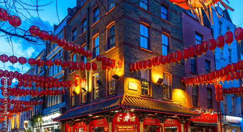 A street corner in Soho's Chinatown with red Chinese lanterns hanging from the walls under a twilight sky and a decorated and ornate typical Chinese restaurant below.