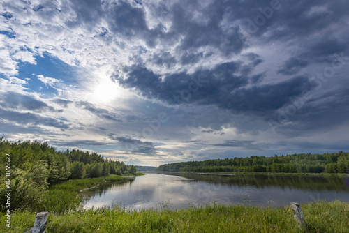 Tranquil river with lush trees, dramatic sky and sun peeking through swirling clouds, creating a picturesque moment in nature.